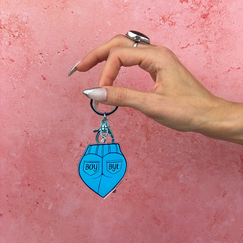 Hand holding a blue heart-shaped keychain with 'BOY' and 'BYE' text on a pink background