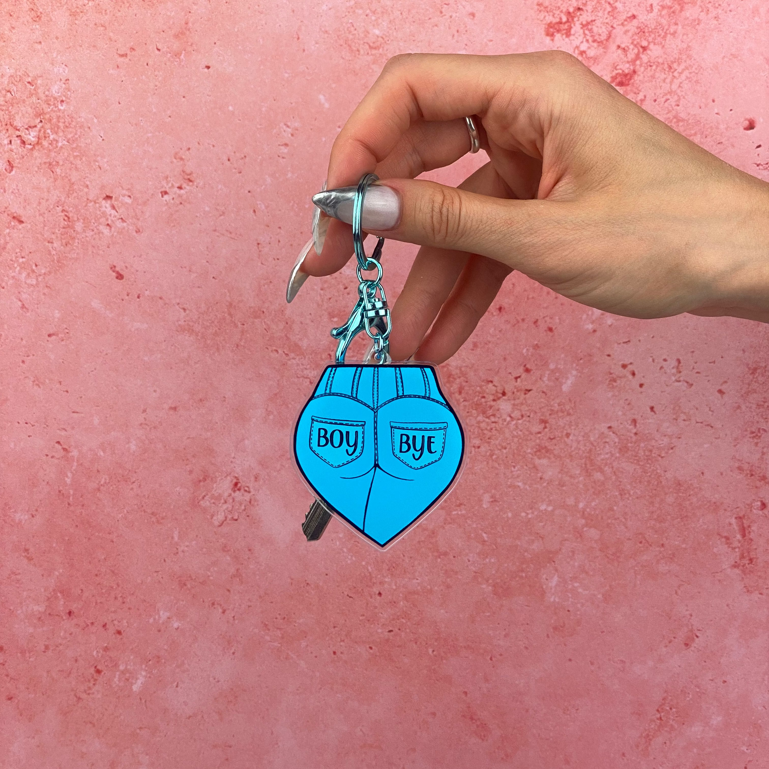 Hand holding a blue heart-shaped keychain with 'BOY' and 'BYT' text against a pink background