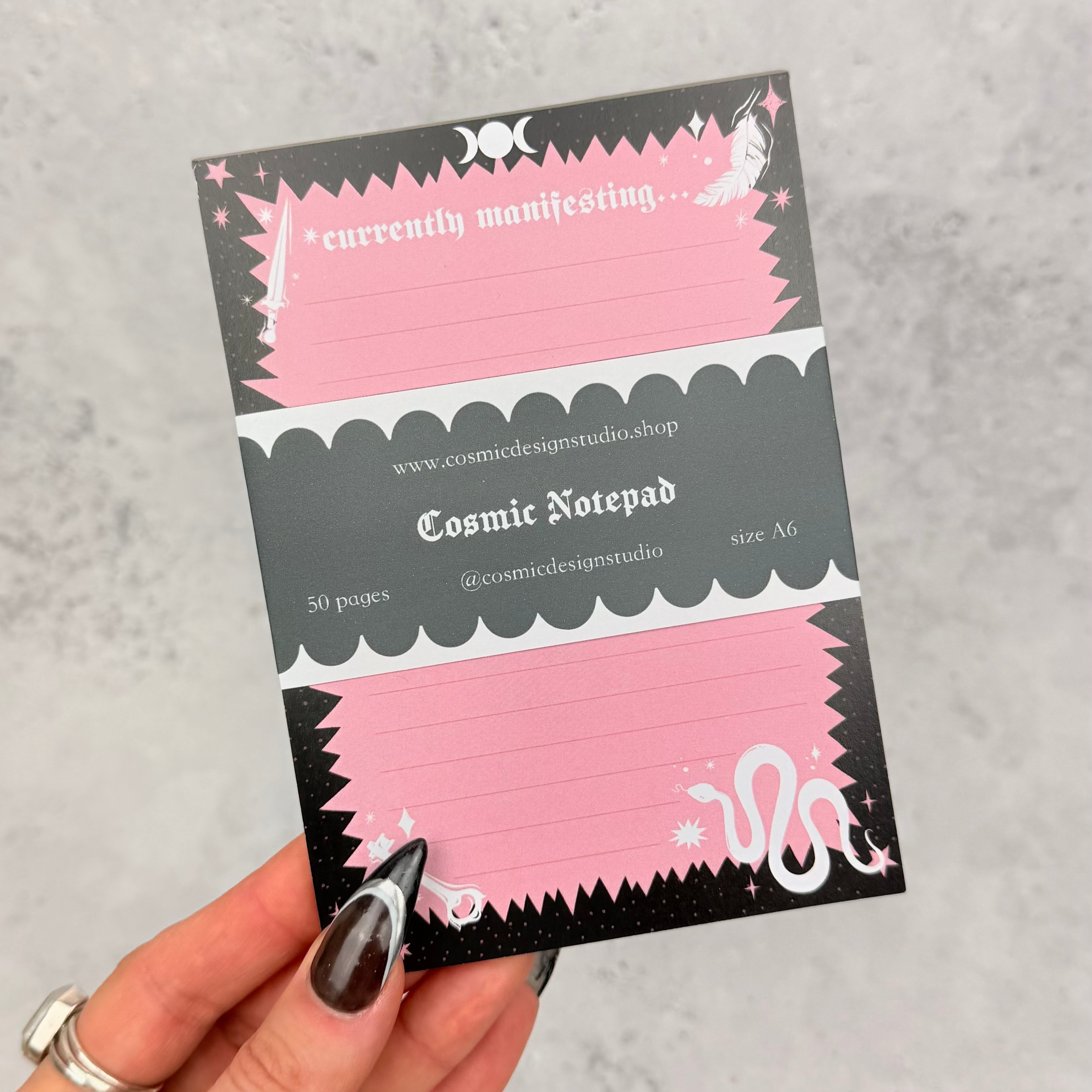 Hand holding a Cosmic Notepad with pink and black design on a gray background