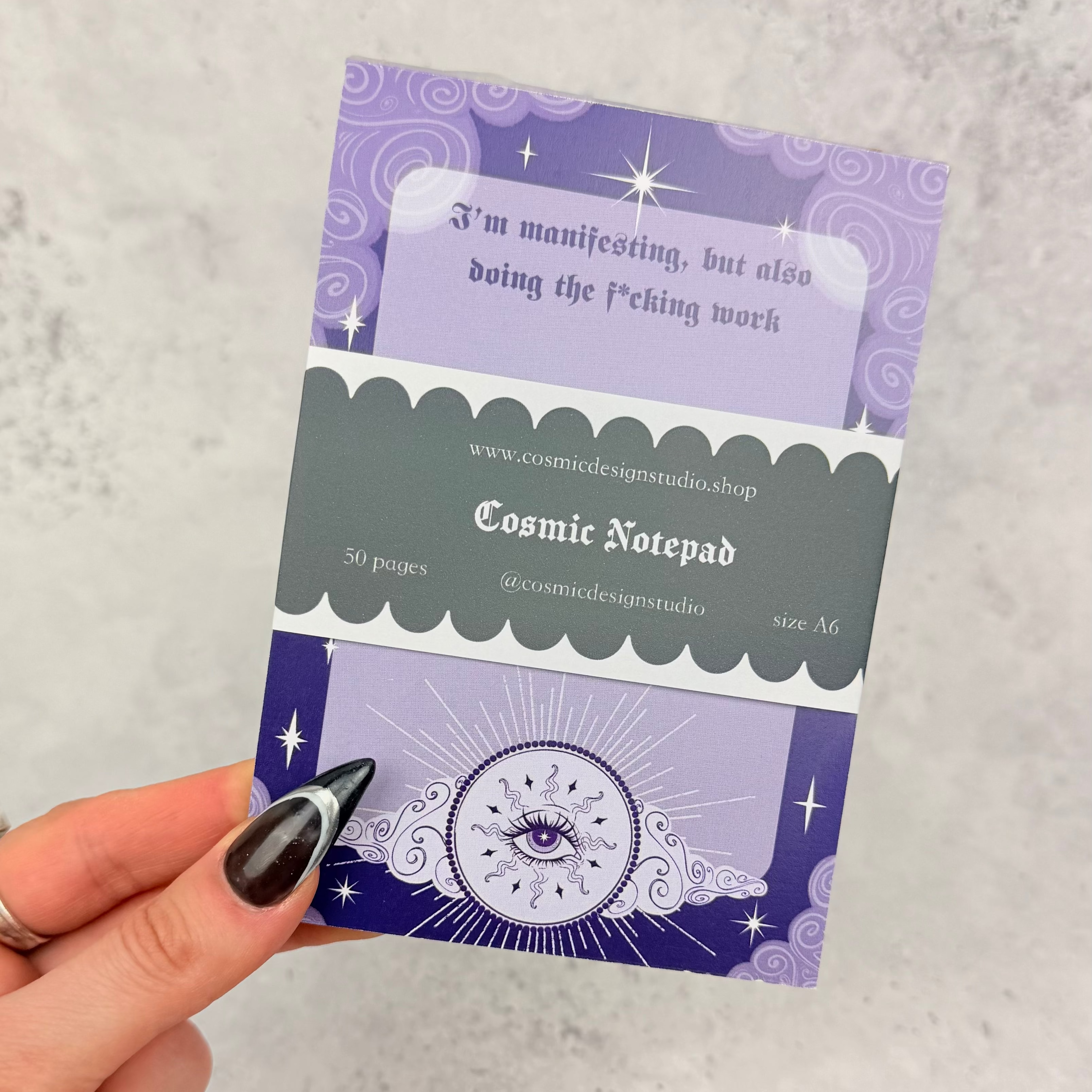 Hand holding a purple and green Cosmic Notepad against a gray background