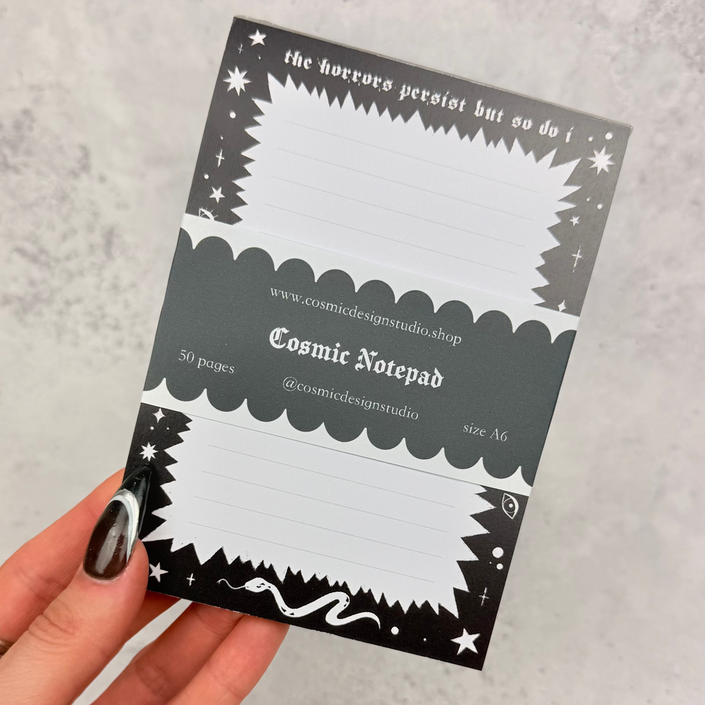 Hand holding a Cosmic Notepad with black and white design on a gray background