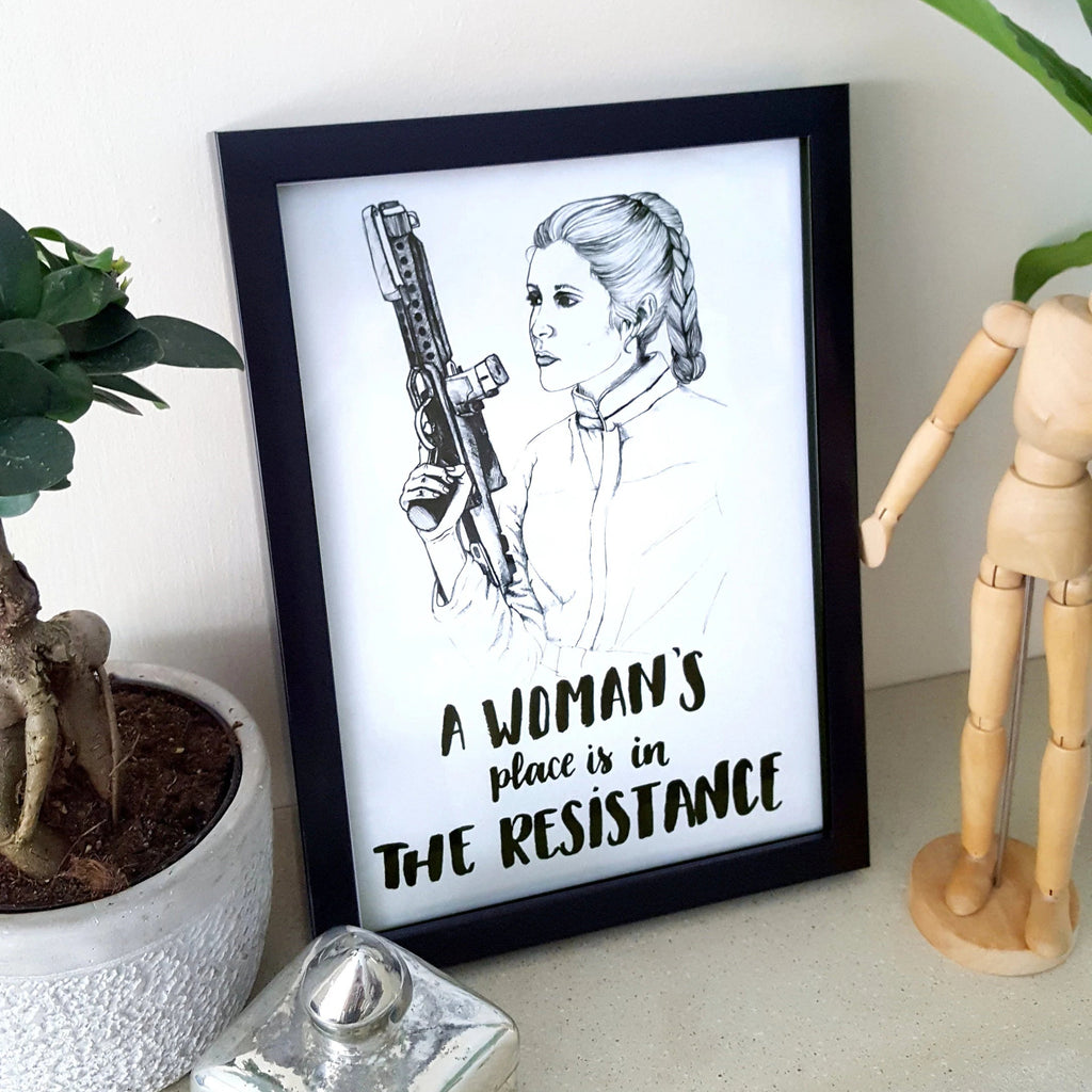 Framed artwork with a woman holding a weapon and text 'A woman's place is in The Resistance' on a white surface with decor items.