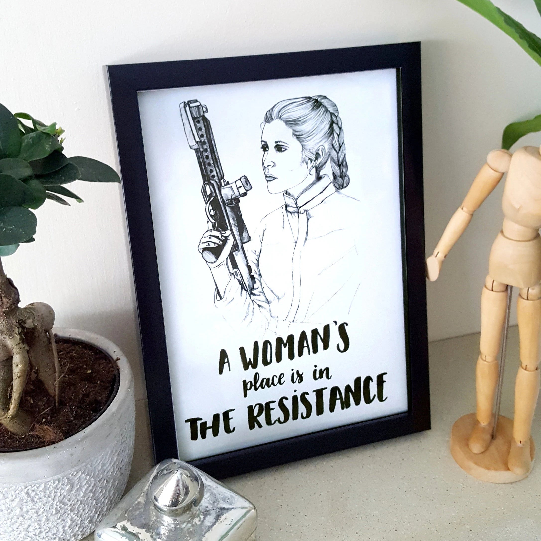 Framed artwork with a woman holding a weapon and text 'A woman's place is in The Resistance' on a white surface with decor items.