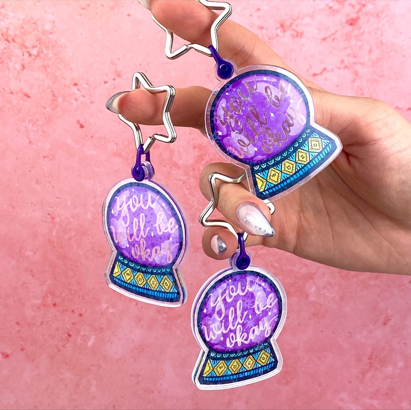 Three star-shaped keychains with purple and blue designs held against a pink background