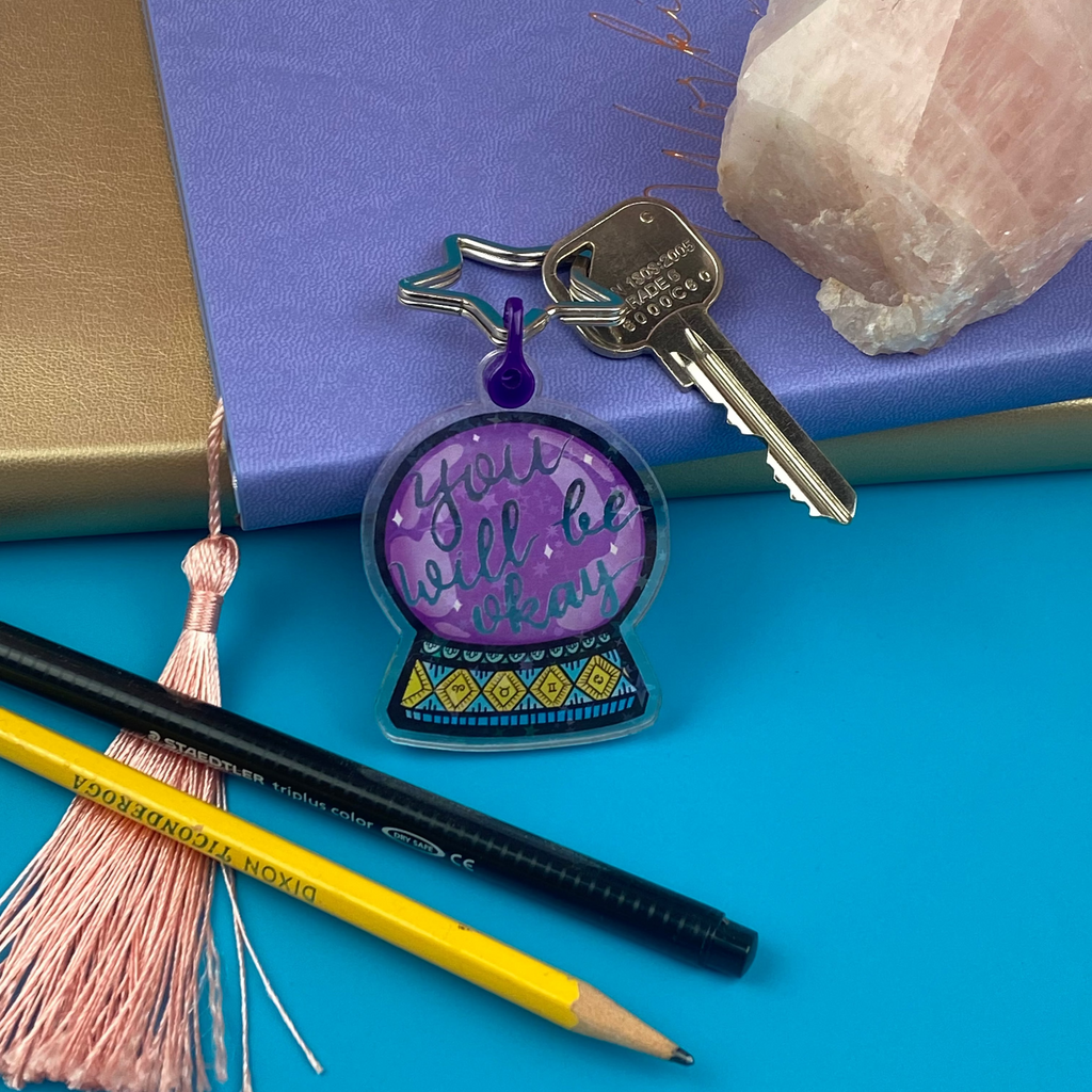 Decorative keychain with purple crystal ball design on a blue surface with pencils and a notebook.